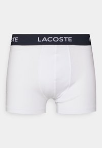 Beli bombažni bokserji z black elastičnim pasom, ki nosi belo "LACOSTE" logotip. Gladka tekstura, prilegajoča zasnova in brez vzorcev.