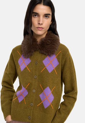 Kobieta w oliwkowozielonym kardiganie z fioletowo-pomarańczowym wzorem w romby oraz brązowym kołnierzem z sztucznego futra, stojąca na białym tle.