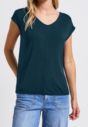 Vrouw draagt een donkerblauwe V-hals top met korte mouwen gecombineerd met lichtblauwe spijkerbroeken, tegen een effen achtergrond.