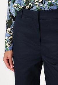 Marineblauwe pantalon met een soepele textuur, voorzakken en subtiele stiksels, gecombineerd met een kleurrijke bloemenhemel met lange mouwen.