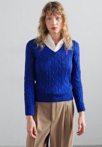 Zils adītas svītras džemperis ar V-veida kaklu un ribotu malu, izgatavots no teksturēta auduma. Uzvilkts pār balto krekli ar apkakli.