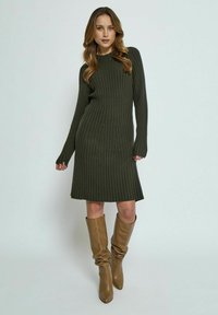 Robe en maille côtelée vert foncé avec un col rond et des manches longues, dotée d'un ourlet évasé, associée à des bottes marron clair jusqu'aux genoux.