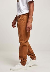 Brune bomuldsbukser med elastiske manchetter, sidelommer og baglommer, samt en afslappet pasform, parret med hvide sneakers og en hvid t-shirt.
