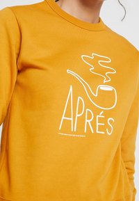Senapsgul sweatshirt med rund halsringning och vit grafik av en rökande pipa samt texten "APRÉS". Mjuk, strukturerad tyg.