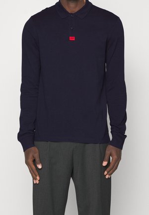 Homme portant un polo à manches longues bleu marine avec un écusson rouge sur la poitrine et un pantalon gris, debout devant un fond uni.