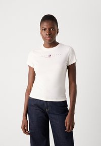 Baltas trumpomis rankovėmis marškinėliai su raudonu ir mėlynu logotipu "TOMMY JEANS NEW YORK CITY" ant krūtinės. Derinami su tamsiai mėlynomis džinsomis.