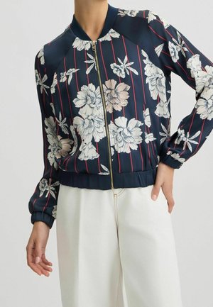 Veste bomber bleu marine avec imprimé floral blanc et fines rayures verticales rouges, associée à un pantalon blanc taille haute, mannequin avec une main sur la hanche.