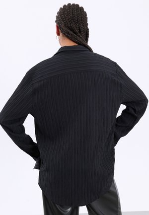 Personne avec des cheveux tressés et des boucles d'oreilles anneaux portant une chemise noire texturée à manches longues et un pantalon noir, de dos, les mains sur les hanches.