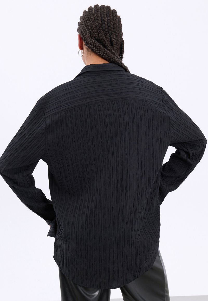 Personne avec des cheveux tressés et des boucles d'oreilles anneaux portant une chemise noire texturée à manches longues et un pantalon noir, de dos, les mains sur les hanches.