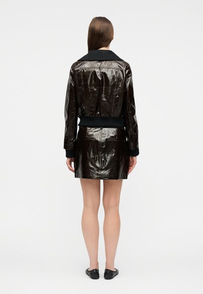 Γυναίκα με την πλάτη γυρισμένη, φοράει γυαλιστερό σκούρο καφέ κομμένο μπουφάν και ταιριαστή μίνι φούστα με μαύρα μοκασίνια σε απλό φόντο.