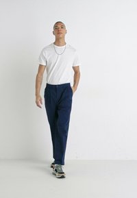 Navy pleated trousers with front pockets, paired with a fitted white t-shirt and multi-colored chunky sneakers. Simple chain necklace detail.
