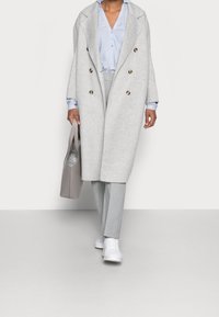 Lysegrå dobbeltradet frakke med store knapper, kombineret med en blå skjorte, grå bukser og hvide sneakers. Holder en grå tote taske.