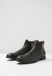 Musta nahkasaappaät nilkkapituiset, joissa on sileä pinta, hopeiset vetoketjut kummallakin puolella ja matala pinohattu. Elegantti ja virheetön muotoilu.