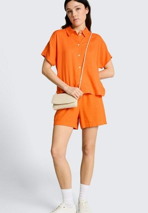 Młoda kobieta w jasnopomarańczowej koszulce z krótkim rękawem i szortach, trzymająca małą beżową torebkę na ramię, stojąca na tle jednolitego tła.