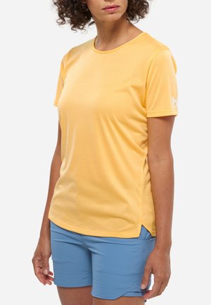 Woman wearing a yellow short-sleeve shirt and blue shorts, standing with arms relaxed by her sides against a plain background.