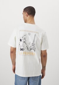 Kortärmad vit bomullströja med grafiktryck på baksidan, föreställande fotbollsspelare och Eiffeltornet, med orange detaljer.