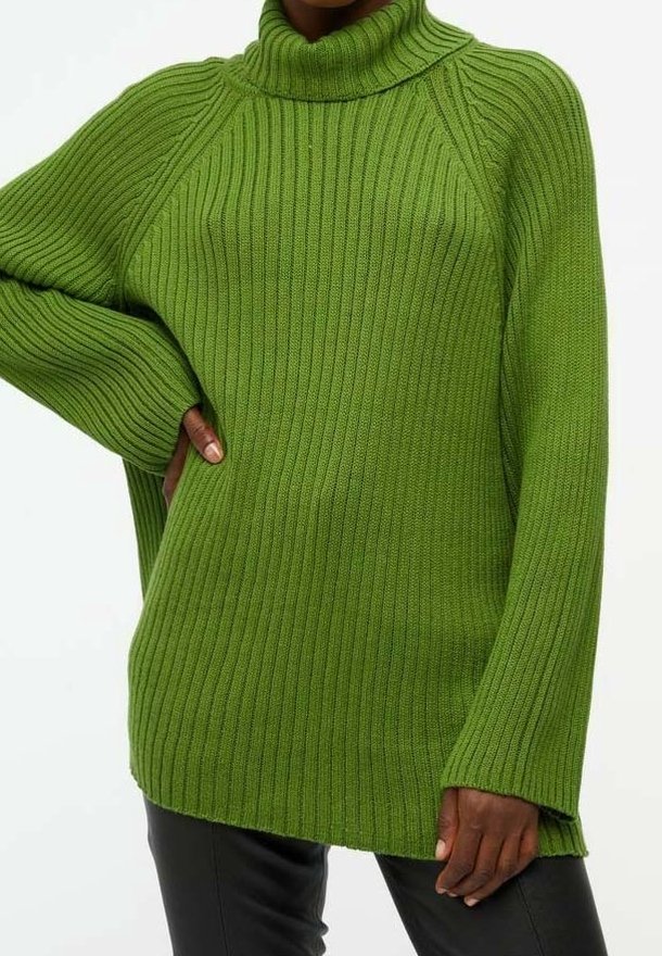 Persoon in een groene geribbelde coltrui met lange mouwen en zwarte broek, hand op de heup.