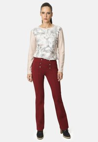 Mujer con el cabello recogido, vistiendo una blusa larga de manga larga de color beige con estampado, pantalones rojos de tiro alto con botones y botas negras hasta el tobillo, de pie.
