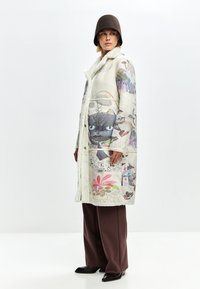 Langer, cremefarbener Mantel mit einem weichen, strukturierten Innenfutter, das farbenfrohe grafische Drucke und Blumenmuster zeigt. Kombiniert mit braunen Hosen und einem Hut.