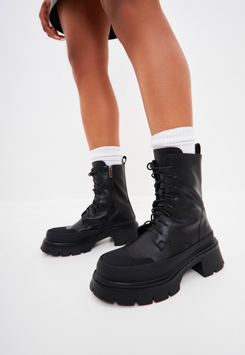 Botas de combate negras hechas de cuero sintético, con una puntera texturizada, cordones en la parte delantera, cremallera lateral y suela gruesa con estrías.