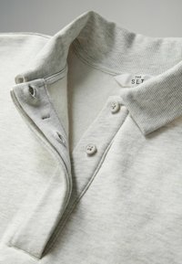 Grijze katoenen polo met een geribde kraag, drie-knopen sluiting en subtiele textuur. Label leest "THE SET." Knopen zijn rond en wit.