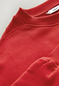 Sweatshirt rouge avec un col et des poignets côtelés, présentant une texture douce et des coutures nettes. L'étiquette sur le col intérieur indique "NEXT."