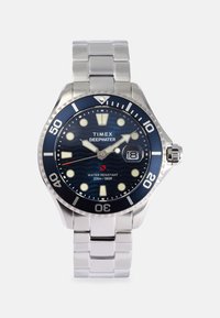 Timex Deepwater óra rozsdamentes acél karkötővel és kék, kerek számlappal, fehér órásjelzőkkel és 3 órakor elhelyezett dátumablakkal.
