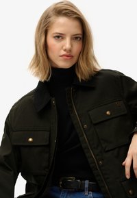 Chaqueta verde oscura con cierre frontal, grandes bolsillos en el pecho y cuello de pana. Llevada sobre un suéter negro de cuello alto, con detalles en dorado.