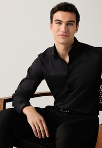 Camicia nera con bottoni, maniche lunghe, tessuto liscio, design aderente e polsini, abbinata a pantaloni neri, indossata da un modello maschile seduto.