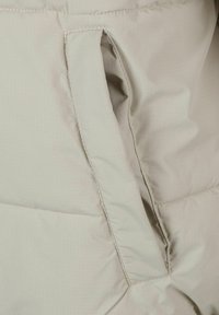 Világos bézs szigetelődzseki anyag, vízszintes varrással, oldalsó zsebnyitással és sima, enyhén fényes felülettel.