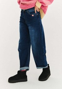 Tmavě modré široké džíny z denimu s ohrnutými nohavicemi a růžovou logo visačkou. Spárováno s černými kotníkovými botami. Texturovaná látka.