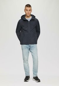 Marineblauer Kapuzenjacke mit einem Frontreißverschluss und zwei Seitentaschen, die eine glatte Textur und ein minimalistisches Design aufweist, kombiniert mit hellen Jeans und Sneakers.
