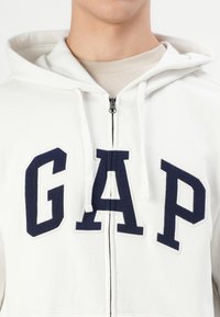 Hvit zip-up hettegenser med mørkeblå "GAP" bokstaver på fronten, snor i hetta og teksturert stoff. Glatt glidelås lukking synlig.