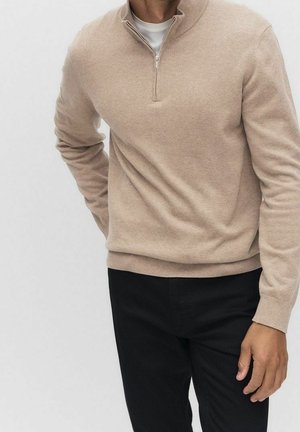 Man draagt beige trui met rits tot op de borst over een wit shirt en zwarte broek, met rechterhand op de heup tegen een effen achtergrond.