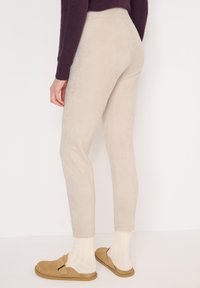 Pantaloni in suede beige con design aderente, vita alta e texture liscia, abbinati a calzini chiari e scarpe marroni senza lacci.