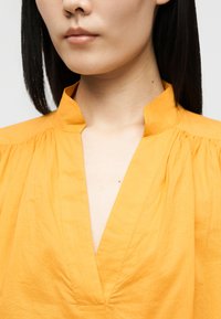 Femme portant une chemise jaune vif à col en V avec un col montant et des épaules froncées, montrée du menton à la poitrine.