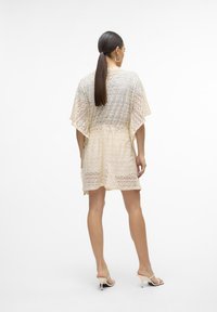 Copertura in crochet beige con motivo a zig-zag, maniche corte e vestibilità ampia, che arriva a metà coscia, indossata con sandali con tacco.