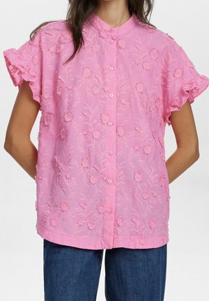 Femme portant un chemisier rose à manches courtes brodé de fleurs, avec des manches à volants et une patte de boutonnage à l’avant, associé à un jean bleu.