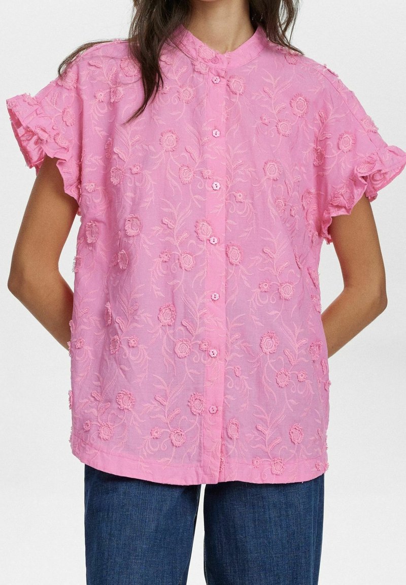 Femme portant un chemisier rose à manches courtes brodé de fleurs, avec des manches à volants et une patte de boutonnage à l’avant, associé à un jean bleu.