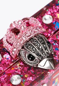 Anello a forma di testa di uccello in argento con corona rosa, adornato di strass. Lo sfondo presenta glitter rosa brillante e grandi gemme trasparenti.