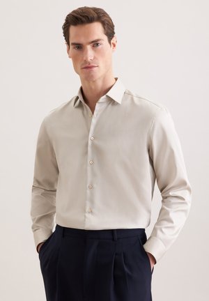 Homme portant une chemise à manches longues beige clair boutonnée et un pantalon bleu marine foncé, les mains dans les poches, sur un fond uni.