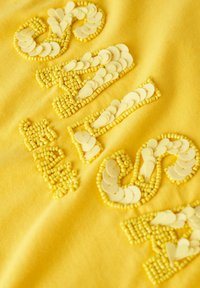 Tela amarilla con letras de aplique hechas de pequeños adornos redondos y en forma de lágrima en tonos amarillos a juego.