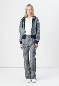Navy tepláková súprava s geometrickým vzorom; zipsová bunda s kapucňou, rebrované manžety; zodpovedajúce nohavice; biely henley tričko; topánky s gumovou podrážkou.