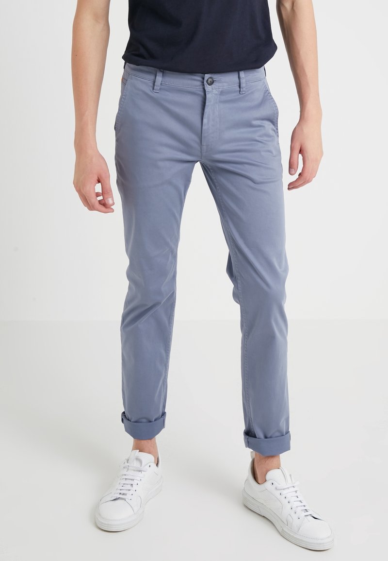Homme portant un pantalon bleu clair retroussé, des baskets blanches et une chemise noire, debout sur un fond blanc uni.