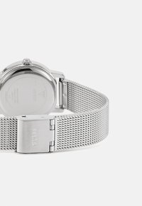 La montre bracelet en maille argentée présente un boîtier métallique rond, avec un design minimaliste et un fermoir marqué "GUESS" sur la sangle.
