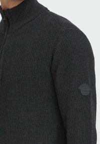 Maglione zip-up in tessuto a maglia nera, con colletto alto e una toppa rotonda sul braccio superiore che recita "Wear the Code Jeanscode Est. 1983."