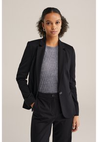 Crni blejzer s izrezanim reverima i prednjim džepovima, uparen s teksturiranom sivoj puloverom. Outfit ima kroj koji prati liniju tijela i dva gumba.