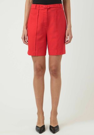 Kvinde iført lyse røde skræddersyede knælange shorts med bæltestropper og sorte spidse hæle, stående mod en ensfarvet baggrund.