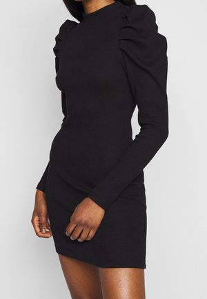 Femme portant une mini-robe noire ajustée à manches longues avec des épaules bouffantes sur un fond clair uni.
