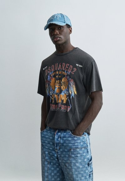 Jeune homme portant une casquette bleue usée, un t-shirt d'entraînement noir à motif graphique et un jean bleu clair à motifs, les mains dans les poches, debout devant un fond gris.